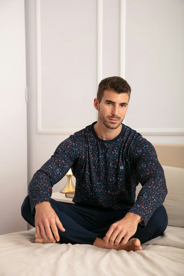Man sitting on a bed wearing a dark blue patterned long-sleeve shirt and dark pants.